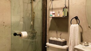 A bathroom with a glass-enclosed shower, a toilet with a basket of toiletries, a shelf with personal care items, and a white towel hanging on a ring.