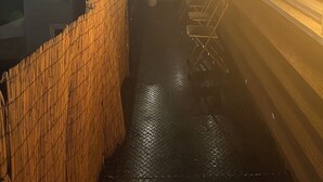 Un pasillo estrecho con una cerca de bambú, barandales metálicos y una fila de sillas.