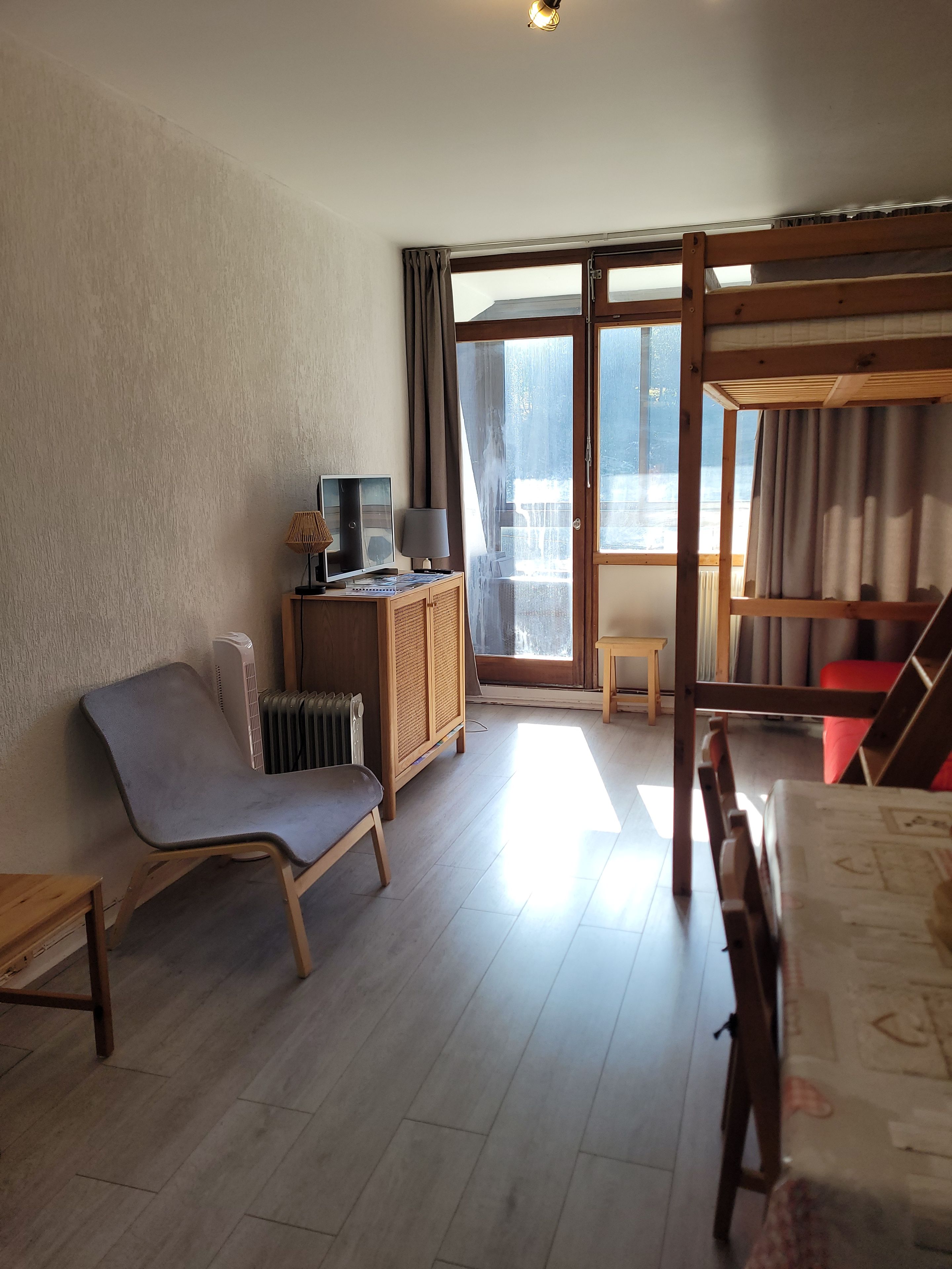Una habitación con una cama nido de madera, una silla, una mesita pequeña, un radiador y una ventana con cortinas.