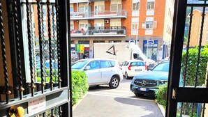 Une grille métallique ouverte donne accès à un parking où se trouvent des voitures, et on aperçoit un bâtiment au fond.