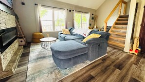 Ein Wohnzimmer mit einer Couch, einem Hocker und einem kleinen runden Tisch. Rechts ist eine Treppe und ein Fenster mit Vorhängen zu sehen.