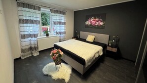 Un dormitorio moderno con una cama grande, una mesita de noche, un radiador, una ventana con cortinas y una lámpara fijada a la pared.