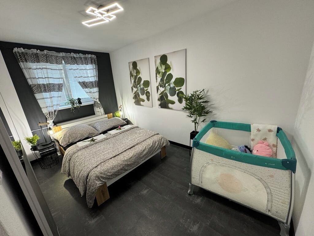 Ein Schlafzimmer mit einem Bett, einem Kinderbett, einem Fenster mit Vorhängen und Wandbildern.
