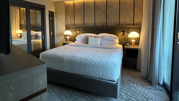 Habitación de hotel con una cama grande, dos mesitas de noche con lámparas, un espejo y un ventanal con cortinas.