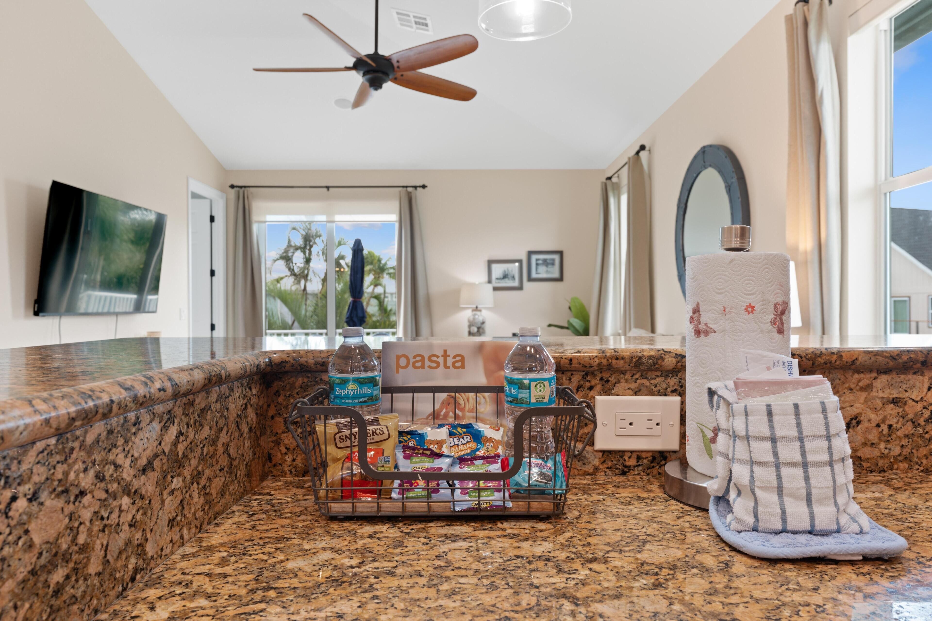 A kitchen with granite countertops, a ceiling fan, a sign labeled 'pasta,' and a basket containing snacks and beverages.