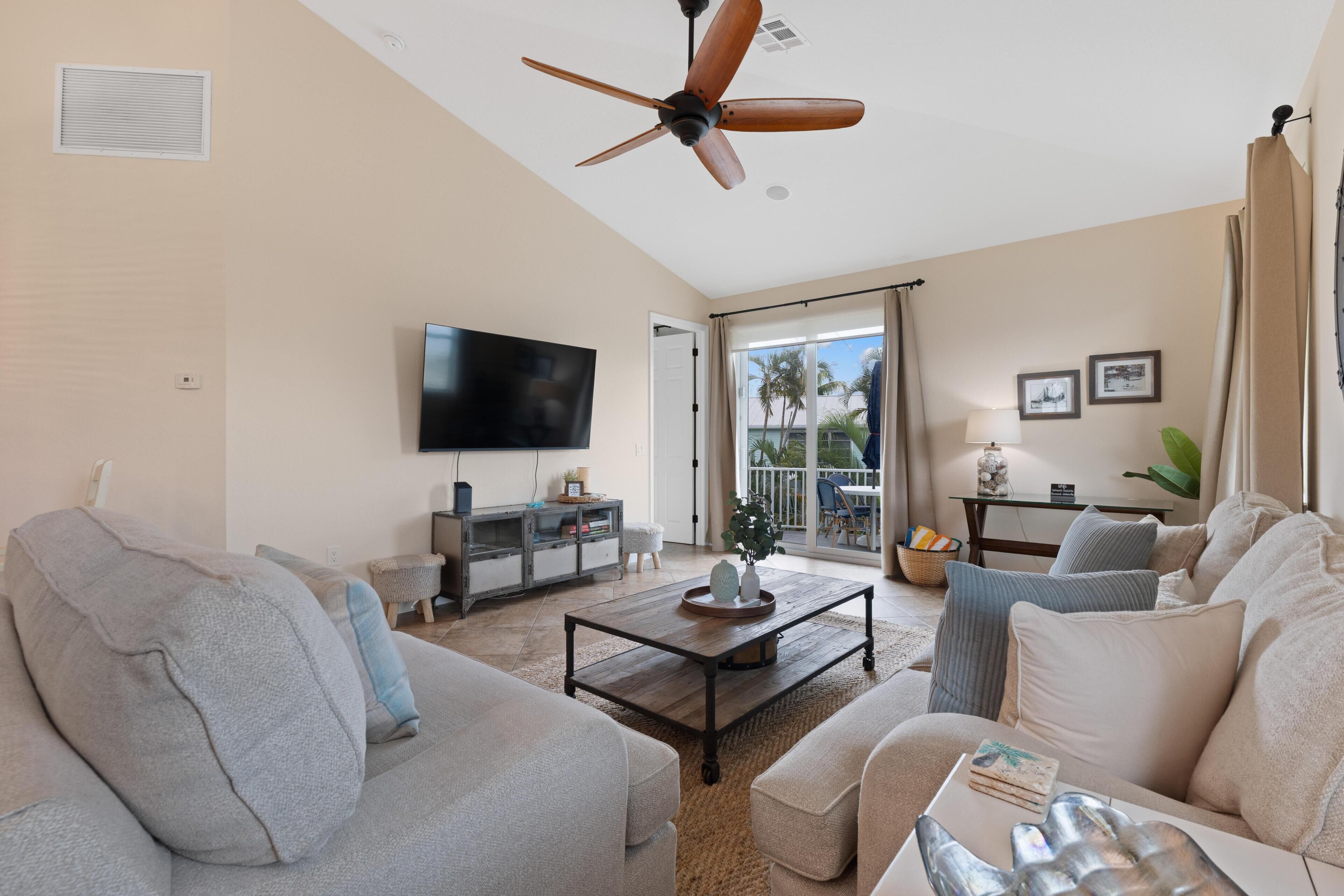 Amplia sala de estar con ventilador de techo, televisor de pantalla plana, sofá, mesa de centro y vistas al exterior.