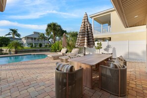 A poolside area with a wooden table and chairs, a striped umbrella, and a view of a residential building. - Sandy Breeze (Fort Myers Beach)
