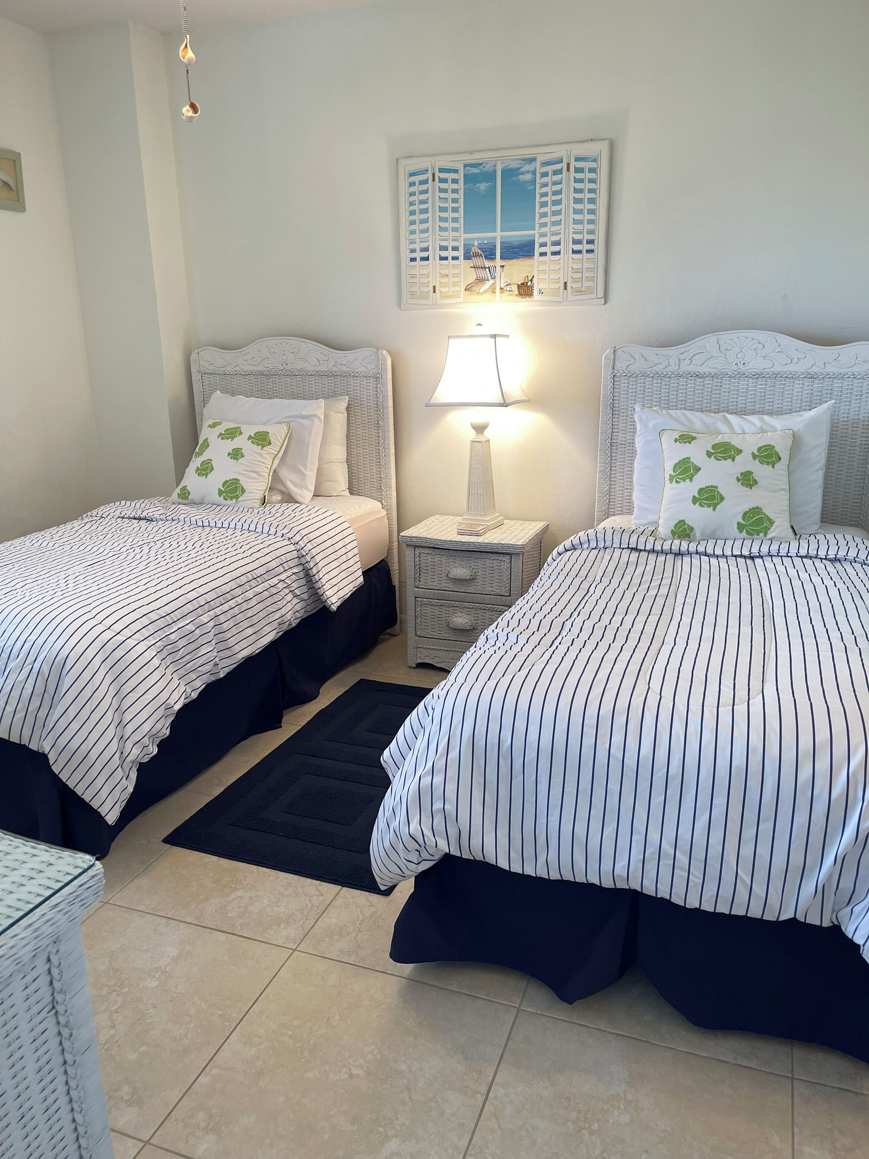 A bedroom with two beds, a nightstand with a lamp, a black rug, and a wall-mounted picture.