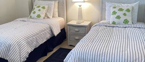 A bedroom with two beds, a nightstand with a lamp, a black rug, and a wall-mounted picture.