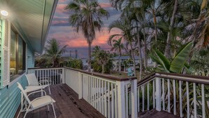A wooden deck with white railings, a couple of white chairs, and a view of palm trees and a sunset. - The Lazy Place (Fort Myers Beach)