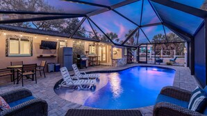 A covered outdoor swimming pool with lounge chairs, a barbecue area, and a TV mounted on the wall. - Happy Daze (Fort Myers Beach)