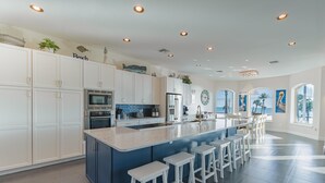 A spacious kitchen with white cabinets, a large island with bar stools, and a view of the ocean through large windows. - Sabal Palms (Fort Myers Beach)