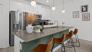 A modern kitchen with white cabinets, stainless steel appliances, a large island with bar stools, pendant lighting, and decorative wall art. - Amazing 4 Bedrooms Home With Pool Close to Disney (Davenport)