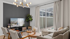 A modern living room with a flat-screen TV, comfortable seating, a decorative plant, and a large window with curtains. - Amazing 4 Bedrooms Home With Pool Close to Disney (Davenport)