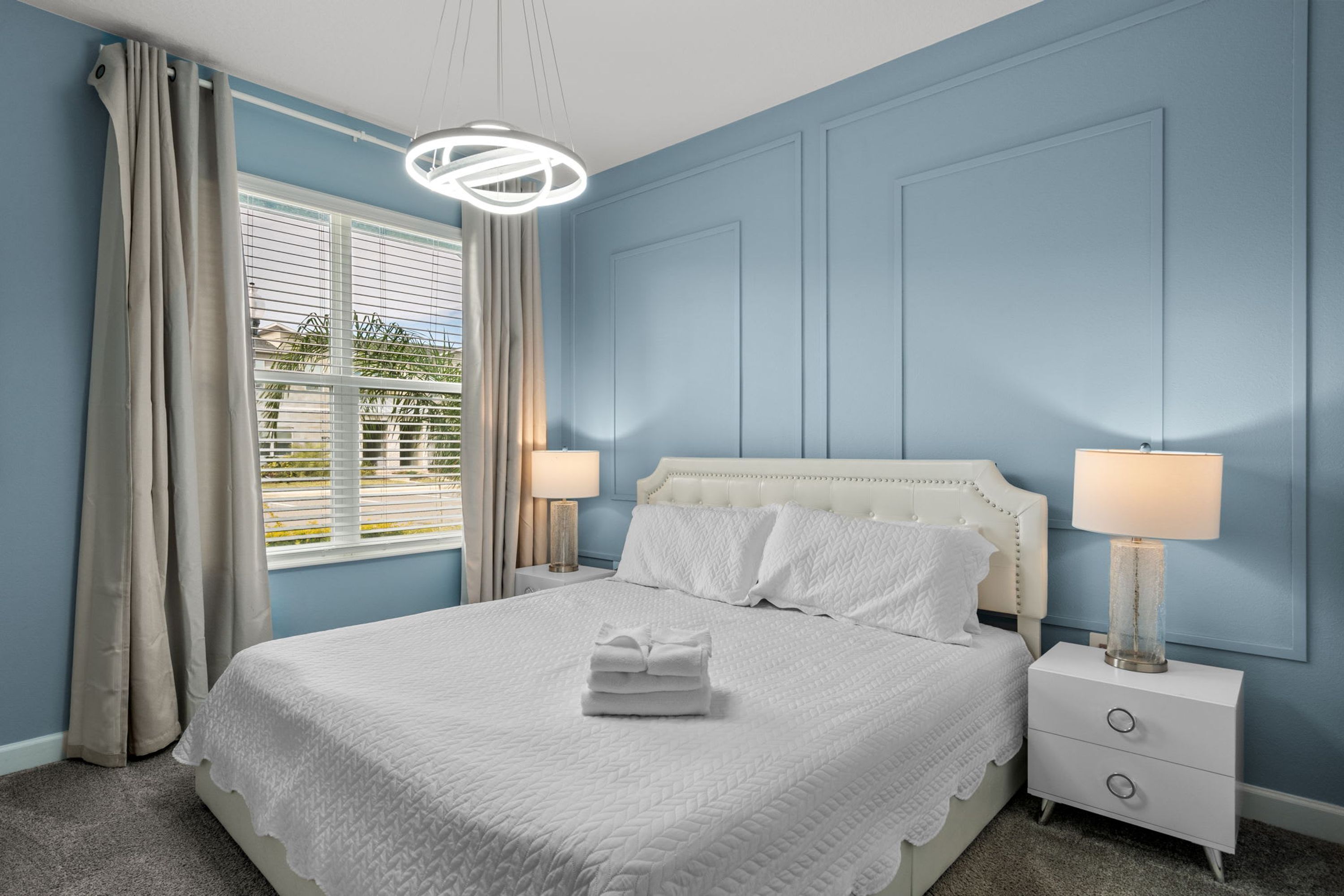 A bedroom with a bed, two bedside tables, a window with curtains, and a hanging light fixture.