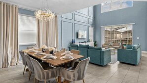 A modern living room with a dining area, featuring a wooden table set for six, grey upholstered sofas, and a large window with blinds. - Luxury 5bed Home With Pool and Game Room (Davenport)