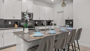 A modern kitchen with white cabinets, a central island, and a dining area with grey chairs. - Luxury 5bed Home With Pool and Game Room (Davenport)