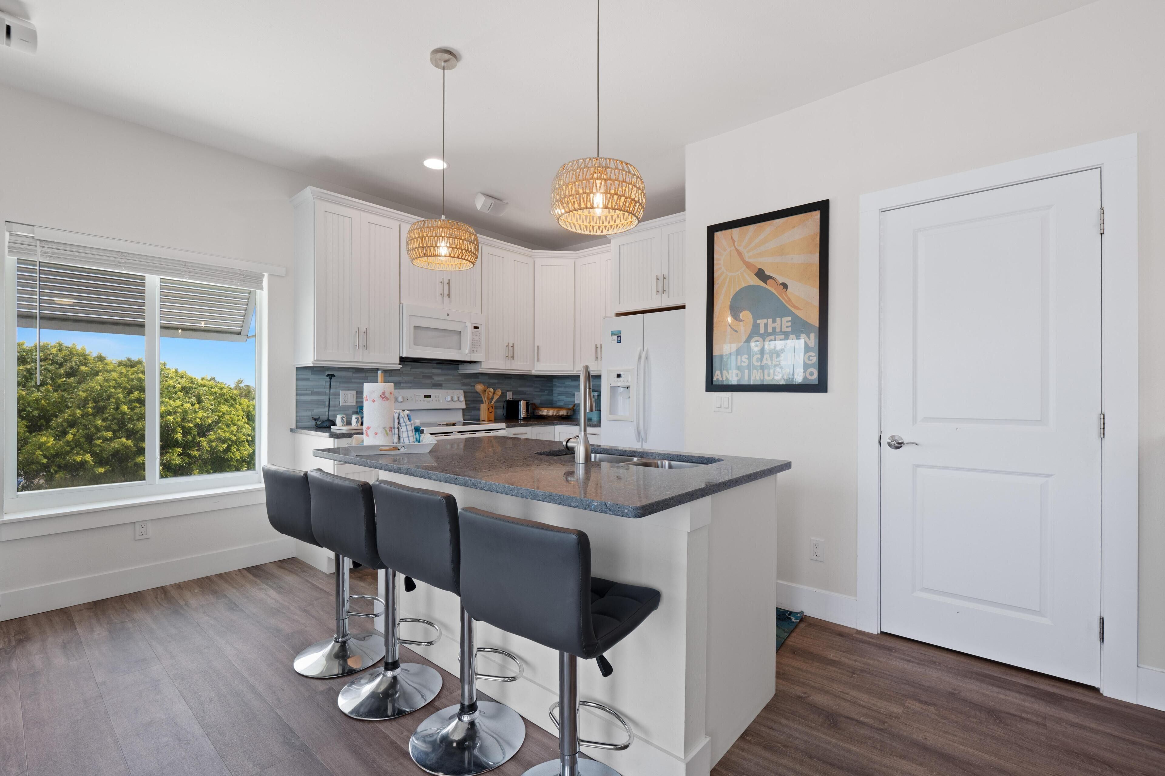 A modern kitchen with white cabinets, a central island with bar stools, pendant lighting, and a view through large windows.