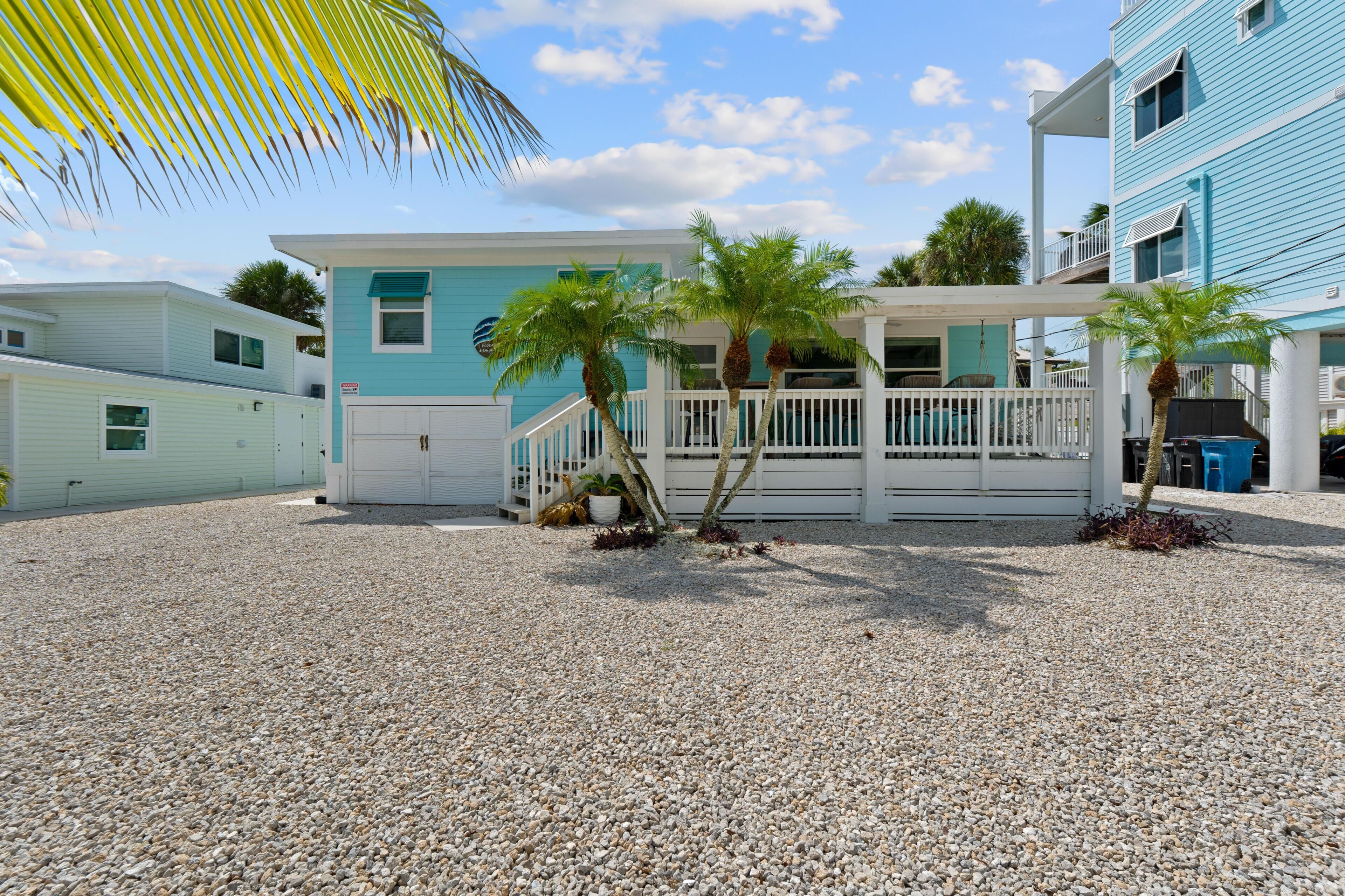 A beachfront property with a covered patio, palm trees, and a gravel driveway.