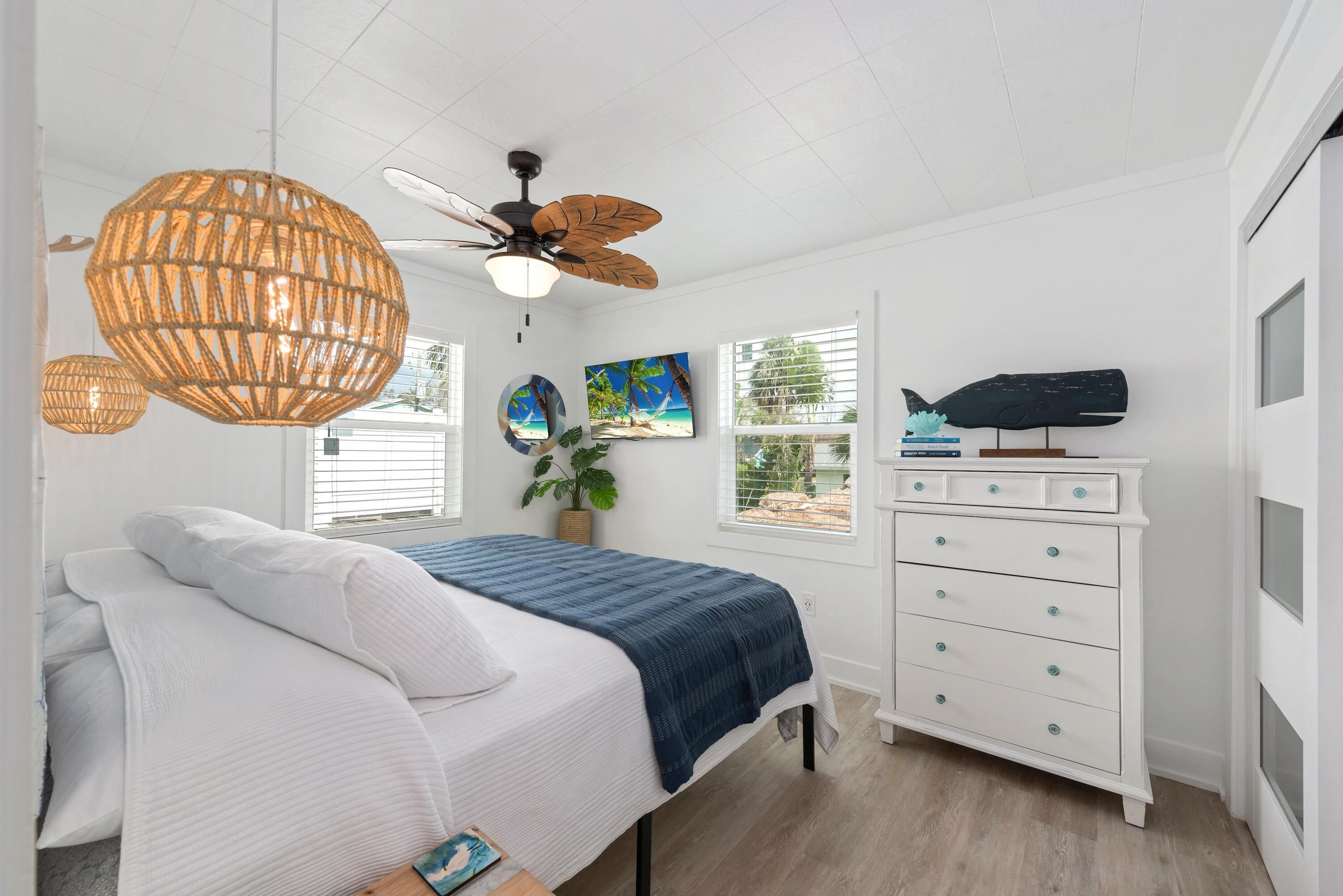 A bedroom with a bed, a dresser, a ceiling fan, and a window with a view of palm trees.