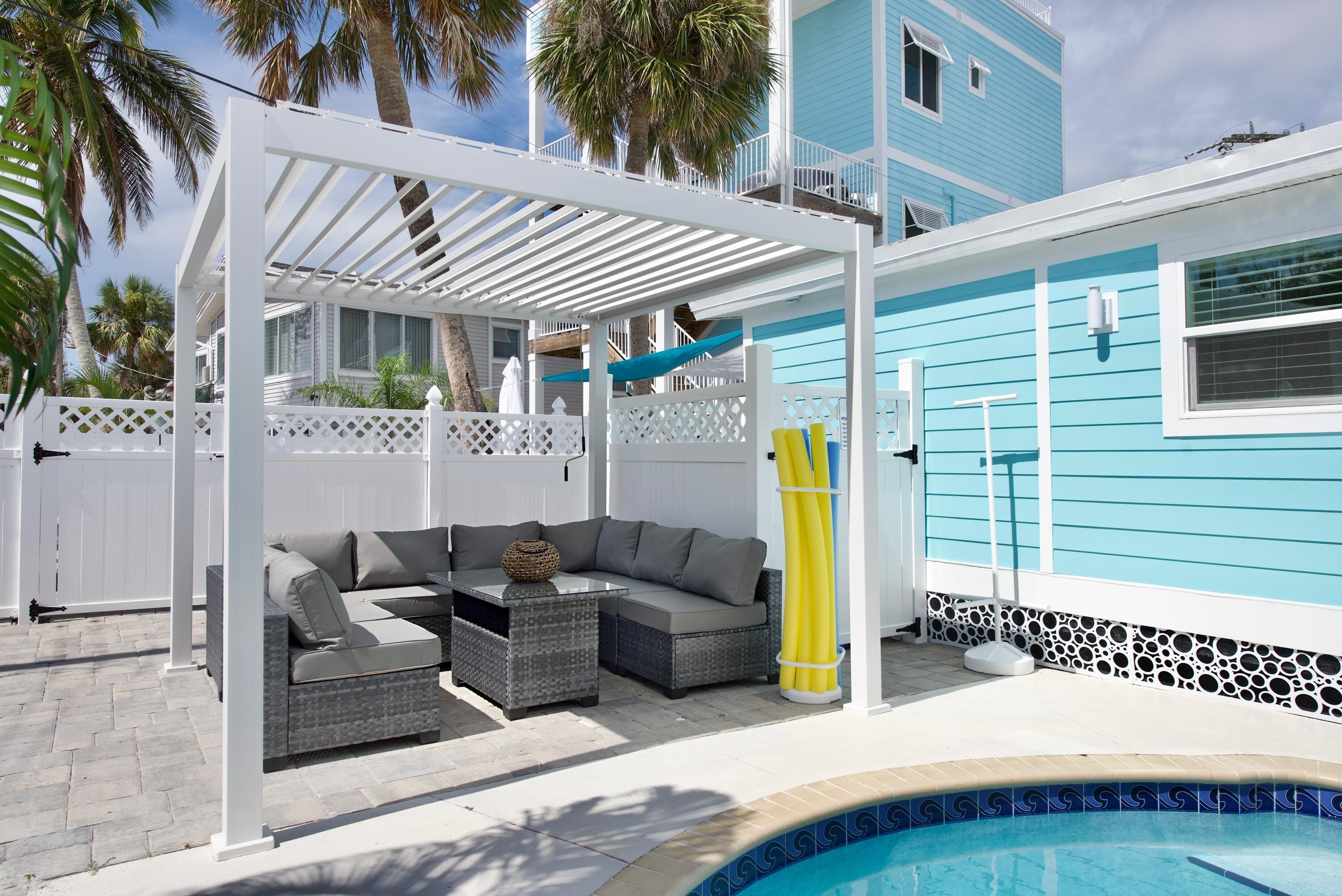 A covered outdoor seating area with a pool, white lattice fence, and a blue building in the background.