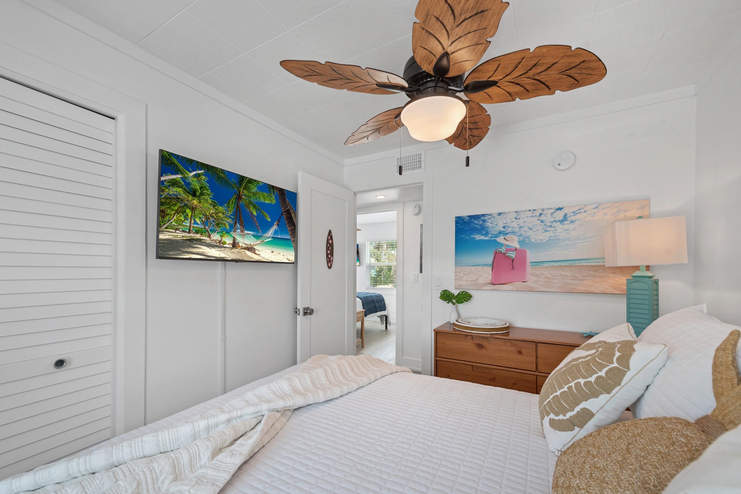 Un dormitorio con una cama, un ventilador de techo, un cuadro de una playa, una mesita de noche y una lámpara.