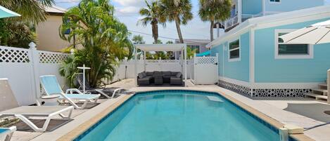 A swimming pool with lounge chairs, a covered seating area, and a white fence.