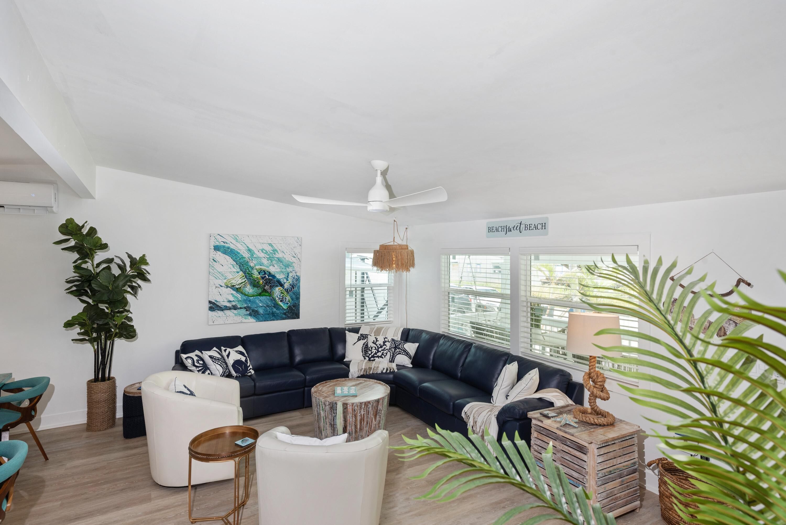 A modern living room with a dark leather sectional sofa, white armchairs, a round coffee table, and a ceiling fan. There is a large window with blinds, a painting on the wall, and a sign that reads "BEACHfront BEACH".