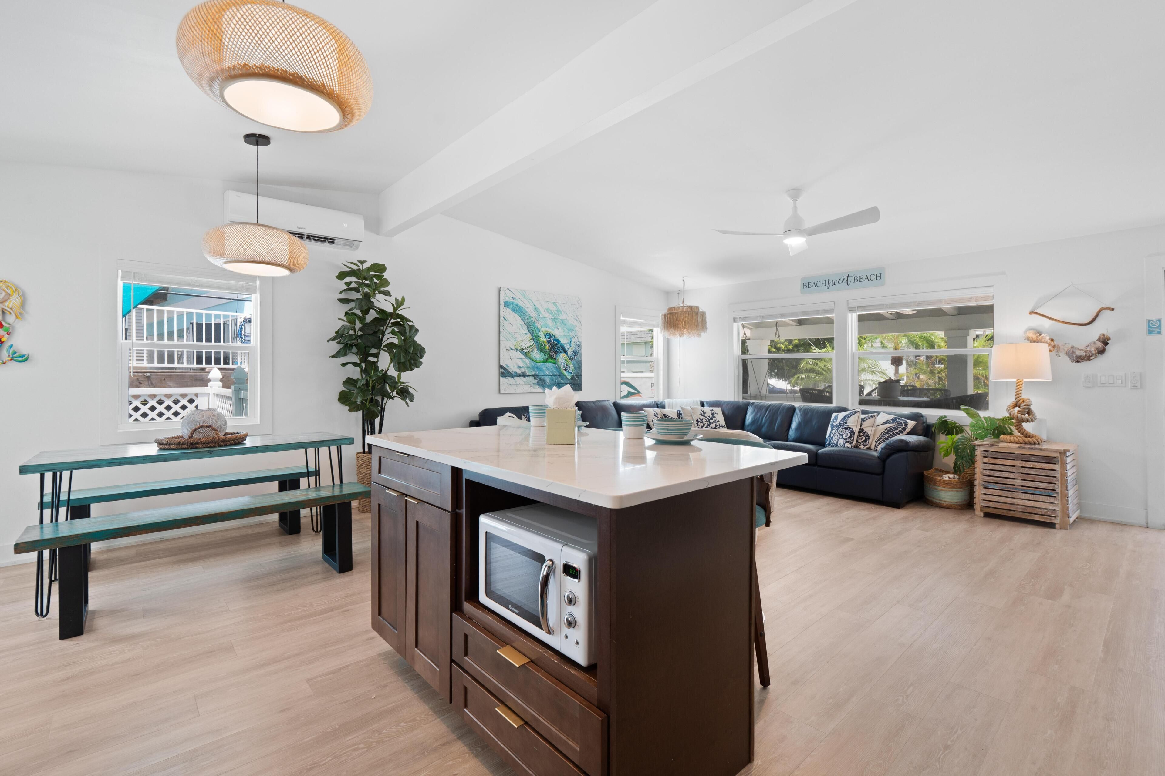 A modern kitchen with a central island, a dining area with benches, a living room with a sofa, and a ceiling fan.