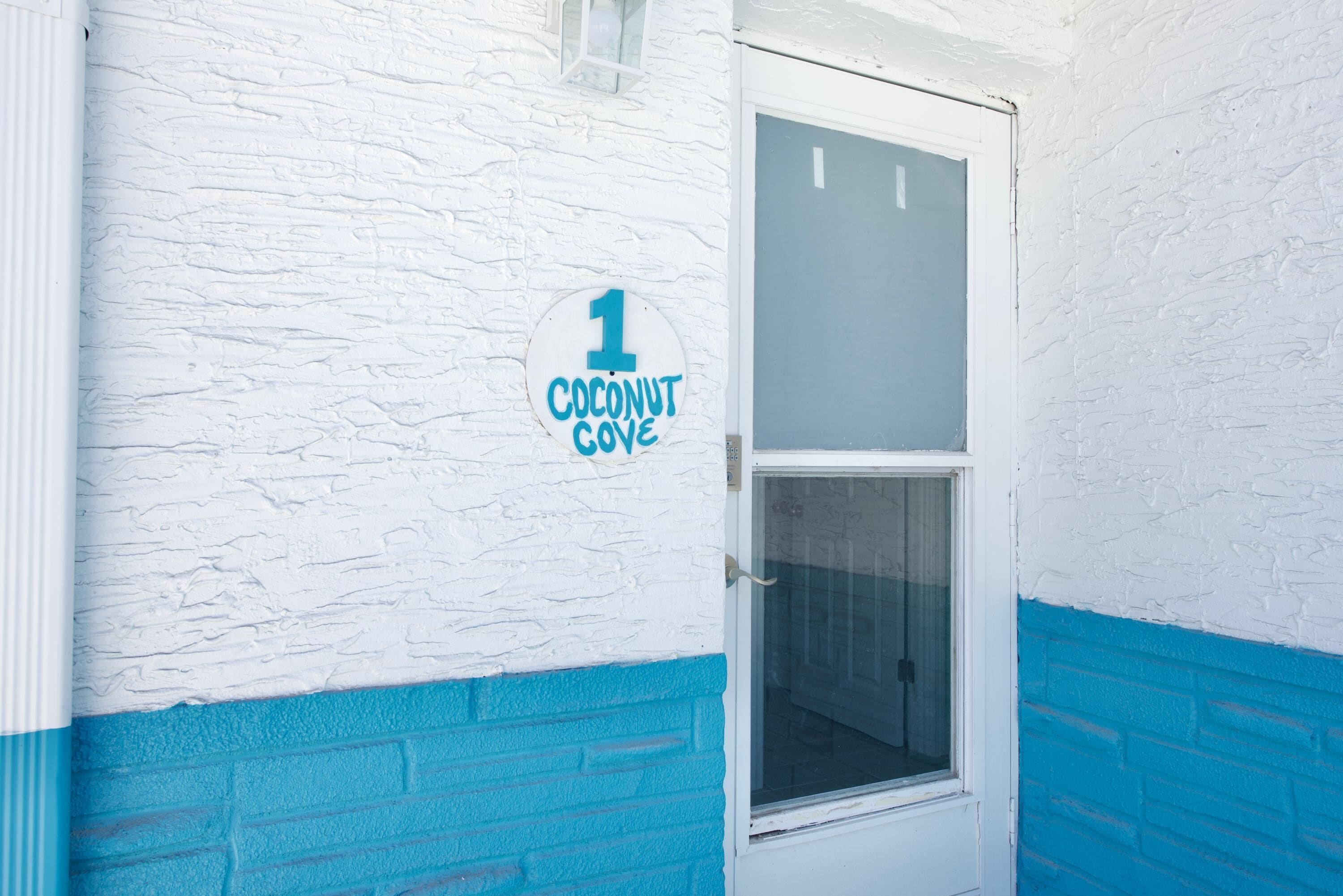 Une porte blanche avec une fenêtre, une partie inférieure bleue et une enseigne indiquant « 1 Coconut Cove ».