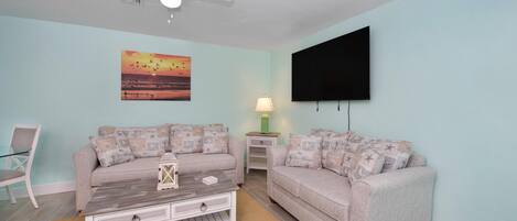 Una sala de estar con dos sofás, una mesa de centro, un televisor, un ventilador de techo y un cuadro.