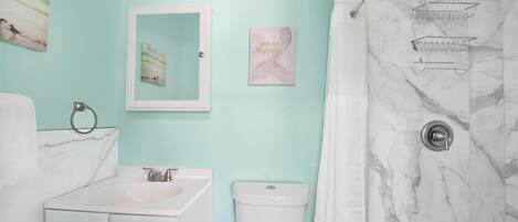 Uma casa de banho com uma sanita, chuveiro e lavatório branco. Possui luzes embutidas na parede, um porta-toalhas e um quadro emoldurado.