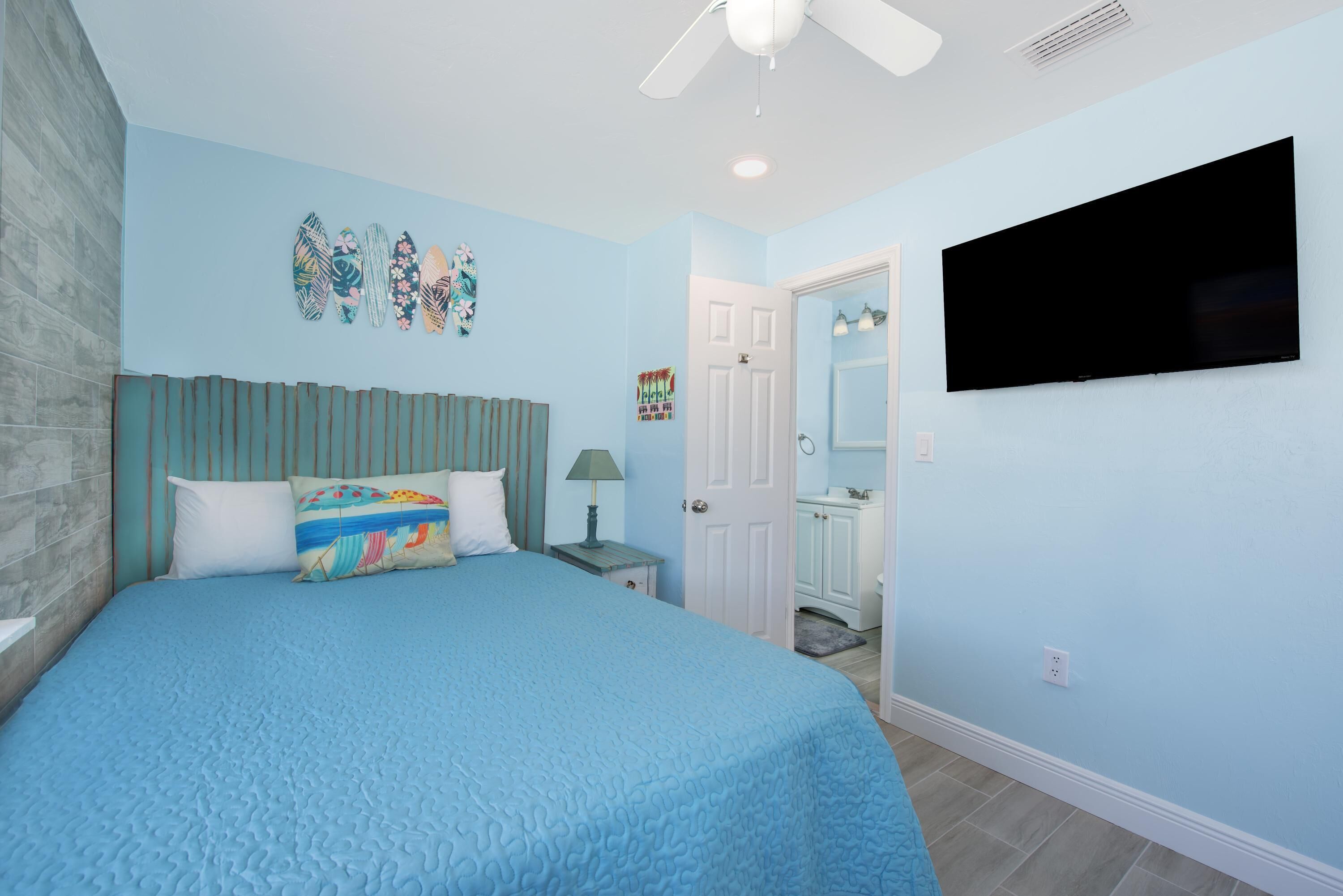 Un dormitorio con una cama grande, un ventilador de techo, un televisor de pantalla plana y una puerta que da al baño.