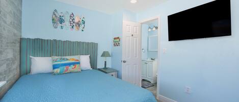 Un dormitorio con una cama grande, un ventilador de techo, un televisor de pantalla plana y una puerta que da al baño.