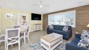 A living room with a blue sofa, white dining table, and chairs, a ceiling fan, and a TV mounted on the wall. - MT Sandy Haven #3 (Fort Myers Beach)