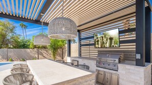 Cocina exterior con encimera de piedra, sillas de mimbre, una parrilla y un televisor montado en la pared.