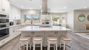 Una cocina moderna con armarios blancos, una isla grande con taburetes, electrodomésticos de acero inoxidable y vistas al exterior.