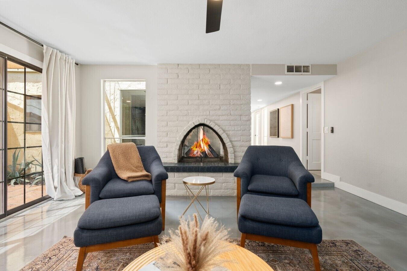 A modern living room with a fireplace, two armchairs, a small round table, and a large window with curtains.