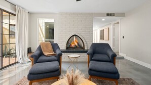 Ein modernes Wohnzimmer mit Kamin, zwei Sesseln, einem kleinen runden Tisch und einem großen Fenster mit Vorhängen.
