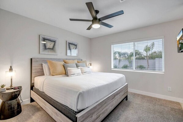 A bedroom with a bed, a ceiling fan, a window with blinds, and two wall-mounted artworks.