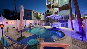 A swimming pool area with lounge chairs and umbrellas, a multi-level building with balconies, and a white picket fence. - Bluewaters Resort (Fort Myers Beach)