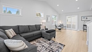Un salon moderne avec un canapé d’angle gris, un pouf noir et un tapis à motifs. Il y a deux fenêtres équipées de stores, une porte et un escalier menant à l’étage supérieur.
