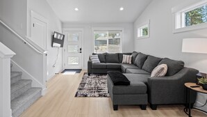 Un salon moderne avec un canapé d’angle gris, un petit pouf, un tapis noir, un escalier et un téléviseur fixé au mur.
