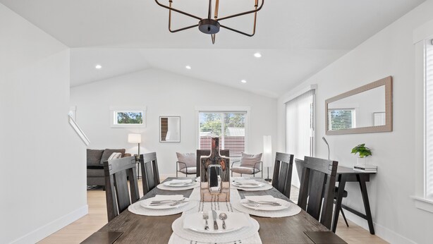 Un coin repas avec une table en bois dressée pour quatre personnes, entourée de chaises sombres, de murs blancs et d’un luminaire au plafond.