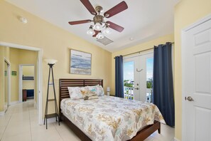A bedroom with a bed, a ceiling fan, a nightstand, a lamp, and a view of the outside. - The Grand Mango (Fort Myers Beach)