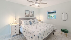 A bedroom with a bed, nightstands, a ceiling fan, a window, and a green stool. - Casa Azul A (Fort Myers Beach)