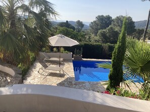 A pool with a stone patio, two lounge chairs, a sun umbrella, and surrounding greenery.
