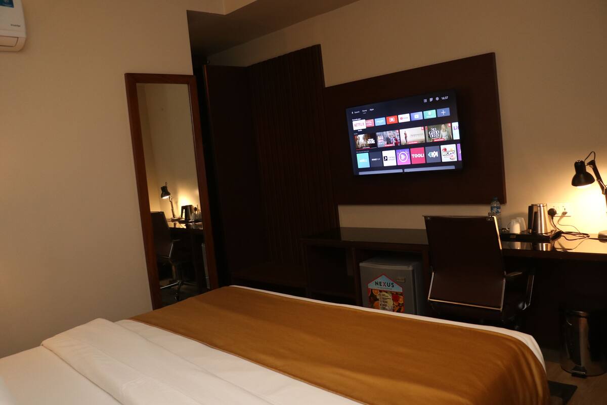 Una habitación de hotel con una cama, un escritorio, un televisor montado en la pared y un refrigerador pequeño con la etiqueta "NEXUS".
