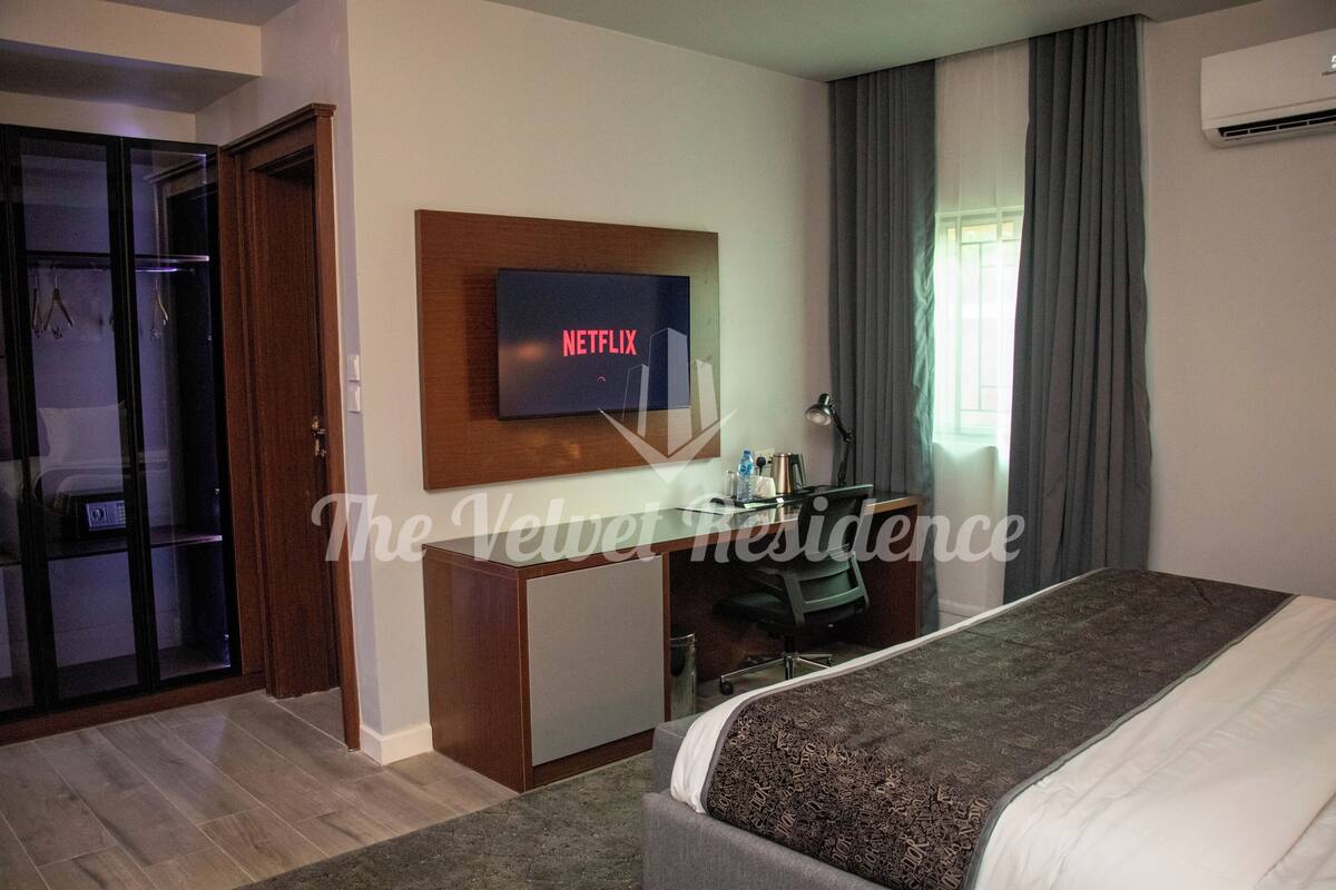 Una habitación de hotel con cama, escritorio con computadora, televisor mostrando 'NETFLIX' y armario.