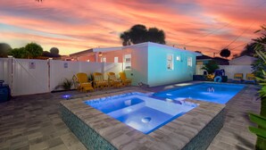 Une piscine avec un jacuzzi, entourée d’un espace délimité par une clôture et agrémentée de chaises jaunes, le tout sous un ciel au coucher du soleil.